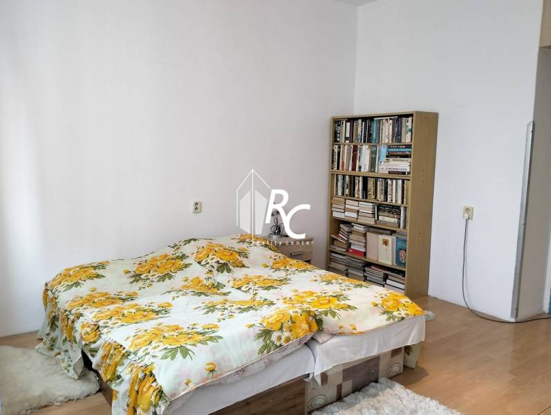 Bedroom in a 2-room apartment with a bed, floral bed linens, and a bookshelf.