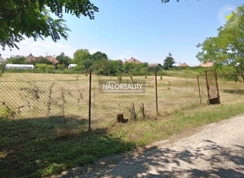 A flat plot of land bordered by a fence in the Gardens of Marcelová, with houses visible in the background.