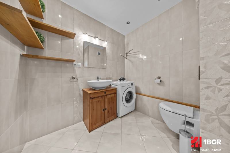 A bathroom in a 3-room apartment with light tiles, wooden shelves, and a washing machine.