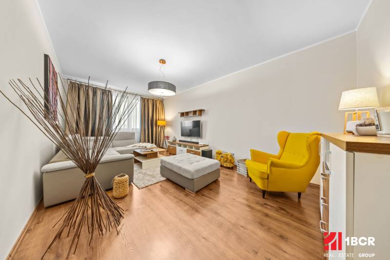 A bright living room in a three-room apartment with wood-patterned flooring and a yellow armchair.