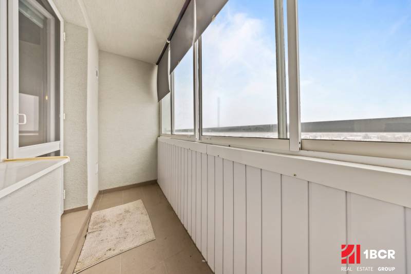 Glazed balcony with brown flooring, blind, and white walls in a 3-room apartment.