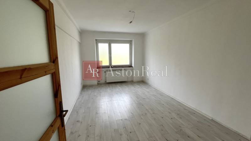 An empty room in a three-room apartment with a wood-patterned floor and a large window.