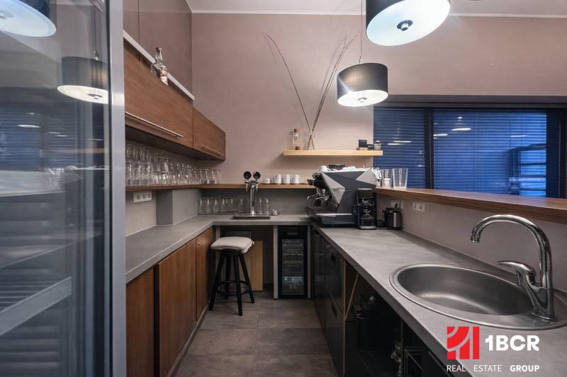 A kitchenette in a commercial space with a coffee machine, wooden cabinets, and a floor with a wooden decor.
