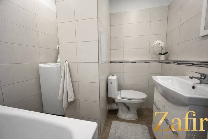 Bathroom in a studio apartment with a bathtub, toilet, and washing machine on a floor with a wooden decor.