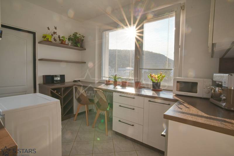 A kitchen with a window, kitchen unit, plants, and a microwave in a 3-room apartment.