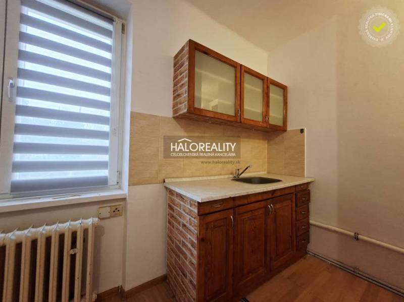 A kitchen in a studio apartment with wooden cabinets and blinds on the window.