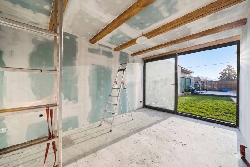 The interior of a family house with a ladder, raw walls, and glass doors to the garden.