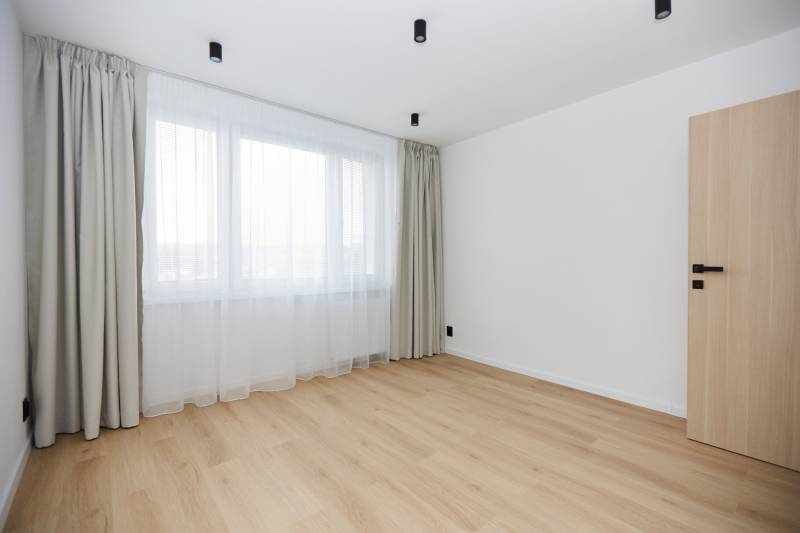 A room in a 3-bedroom apartment with a wooden decor floor and a large window with curtains.
