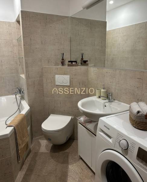 Bathroom in a 2-room apartment with a bathtub, washing machine, and soft beige tiles.