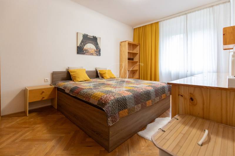 A bright bedroom in a two-room apartment with a bed, yellow pillows, and a wooden decor floor.