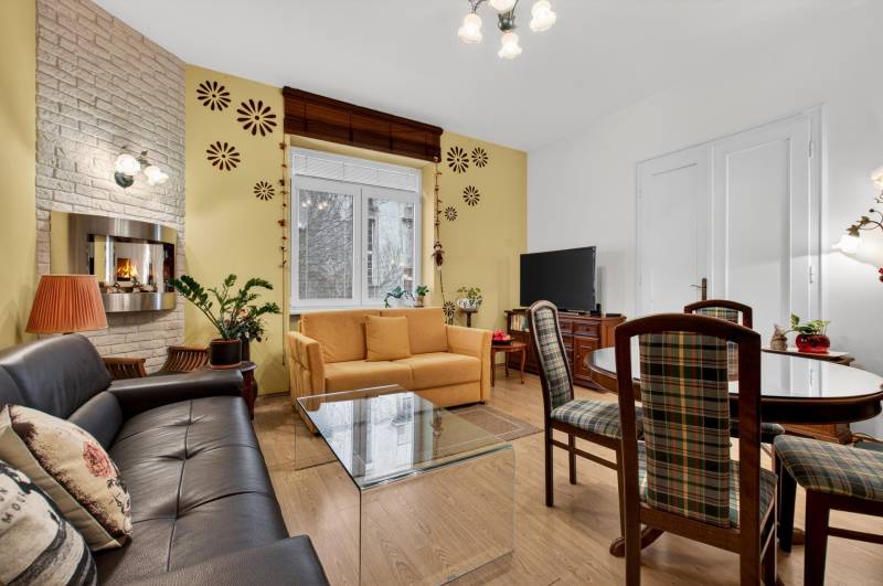 A bright living room with a fireplace, sofa, TV, and wood-patterned flooring in a three-room apartment.