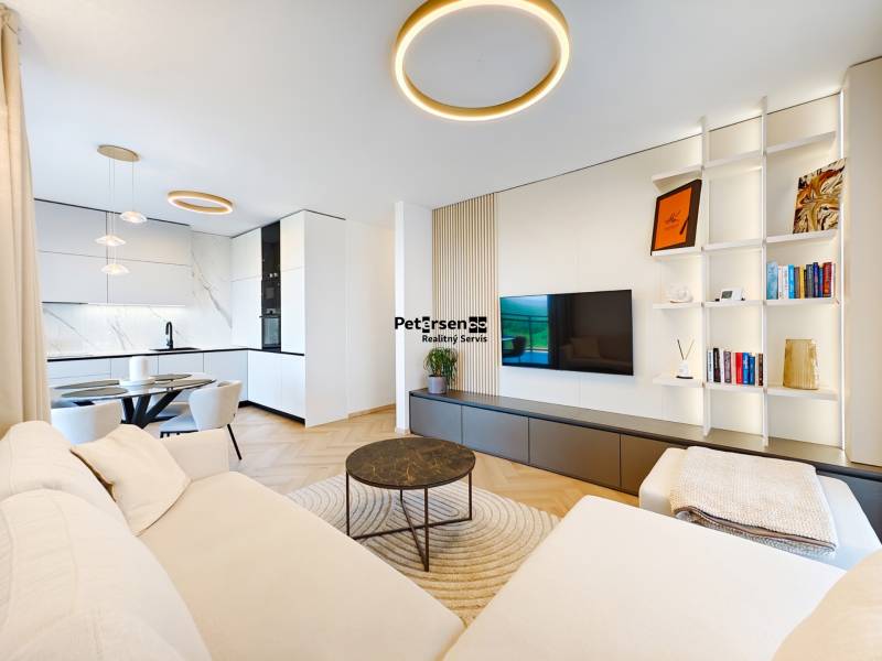 Living room of a two-room apartment with a sofa, kitchen, and wooden decor flooring.