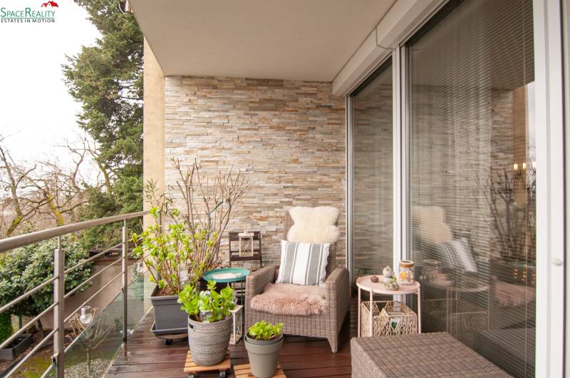Balcony of a 2-room apartment with comfortable seating, flower pots, and glass doors.