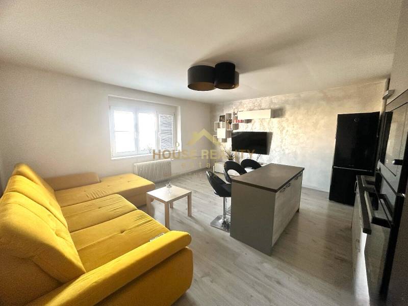 Living room with a large yellow sofa, kitchen island, and television in a 2-room apartment.