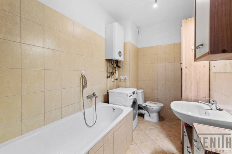 Bathroom with shower, washing machine, and sink in a 2-room apartment with beige tiles.