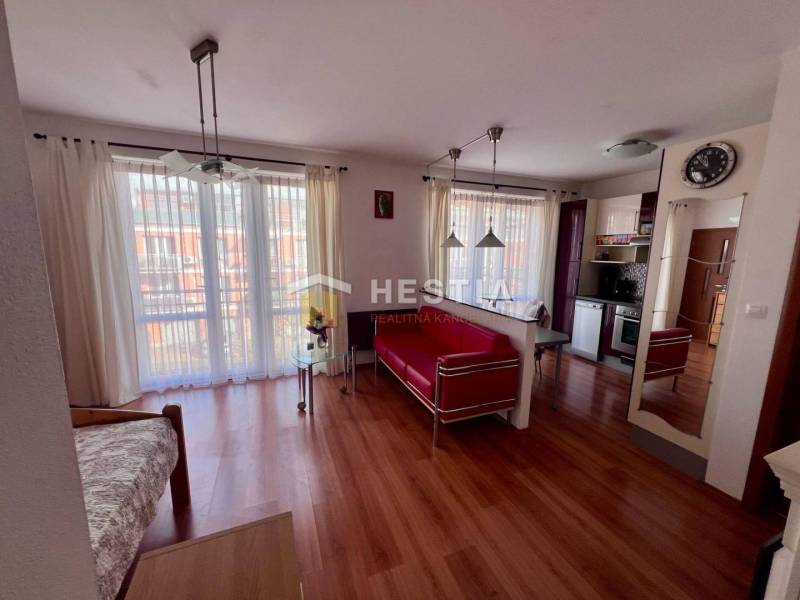 Interior of a 1-room apartment with wooden flooring, a kitchen, light curtains, and comfortable seating.