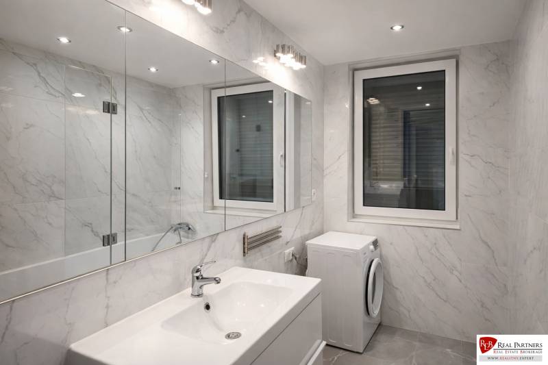 Bathroom in a 2-room apartment with a bathtub, sink, and washing machine, with marble-look tiles.