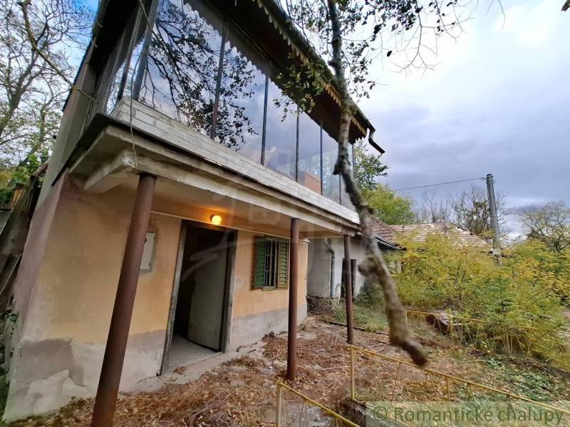 A cottage in Kamenín with a glass-enclosed balcony, surrounding nature, and an overgrown yard.