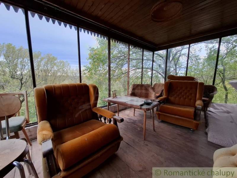 A veranda with chairs and a wooden floor, a cottage surrounded by greenery.