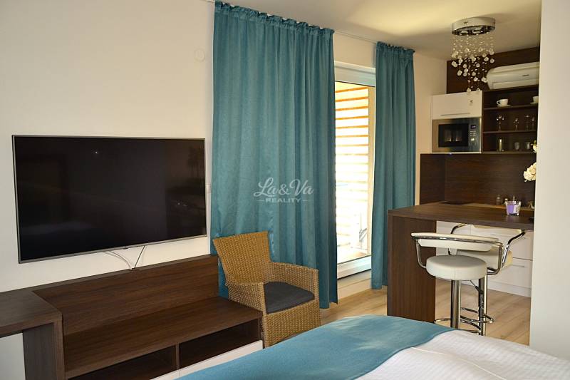 Studio apartment with a TV, wooden furniture, kitchen counter, and turquoise curtains.