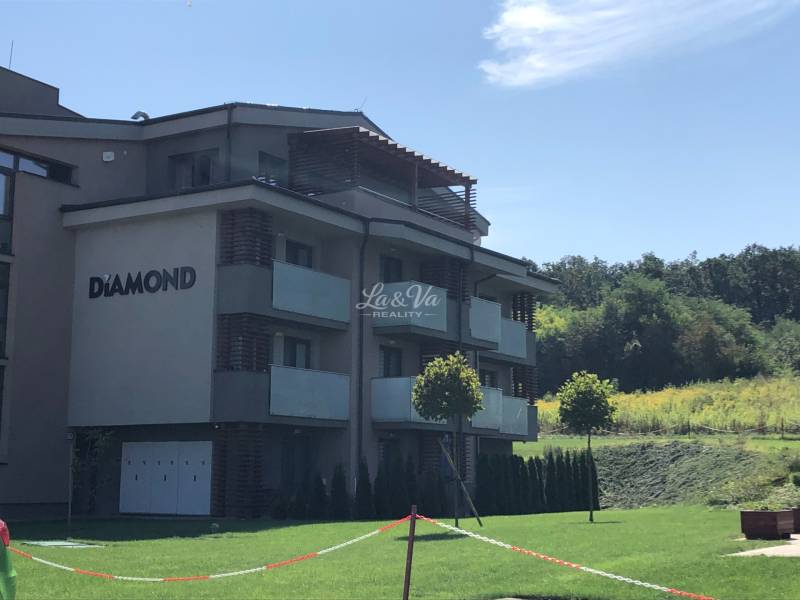 A 2-room apartment building in Podhájska surrounded by greenery and a cloudless sky.