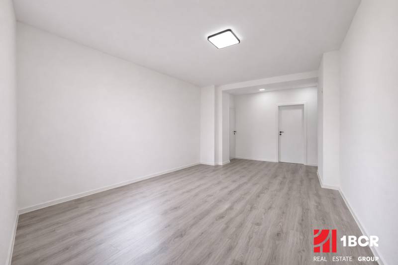 A spacious room in a 3-room apartment with wood-patterned flooring and white walls.