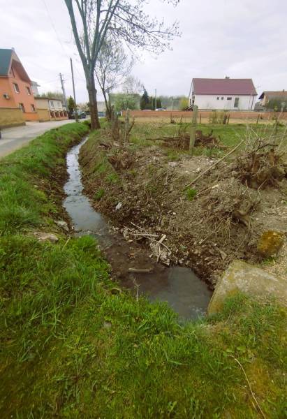 A stream near residential plots in Horná Kráľová surrounded by greenery and trees.
