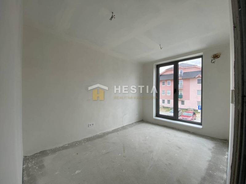 A room in a 2-room apartment with a raw floor and a large window overlooking the street.