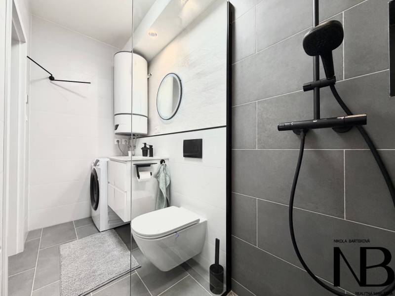Bathroom in a studio apartment with a sink, shower, and washing machine on light-colored tiles.