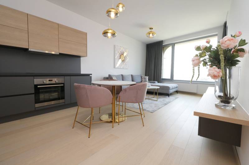 Living room with kitchen, wooden decor flooring, light-colored sofa, dining table, flowers.