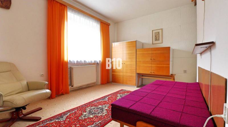 A room with a carpeted floor, a cream armchair, a colorful bed, and a wardrobe in a family house.