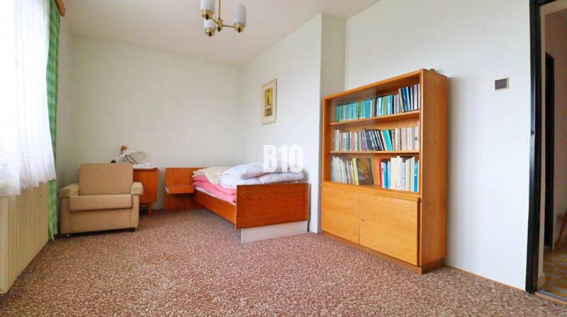 A room in a family house with a bed, a bookshelf, and an armchair on a carpet.