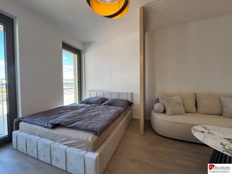Bedroom of a one-room apartment with a bed, sofa, and wooden decor flooring.
