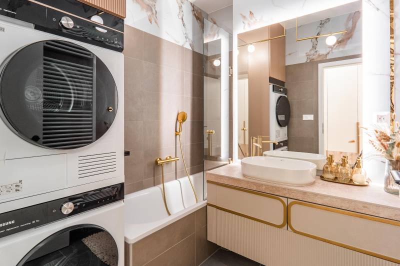 A bathroom in a 3-room apartment with a bathtub, sink, dryer, and elegant gold accessories.