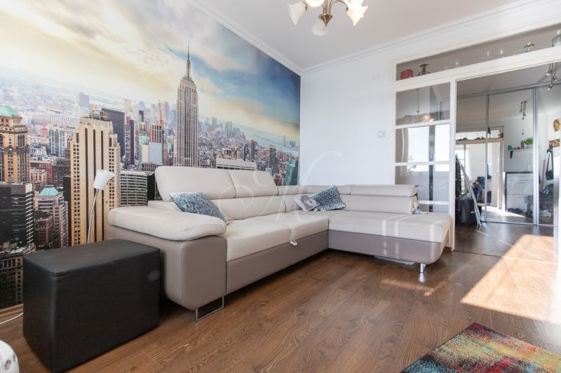 Living room in a 2-room apartment with a beige sofa, city photo wallpaper, wooden decor flooring.