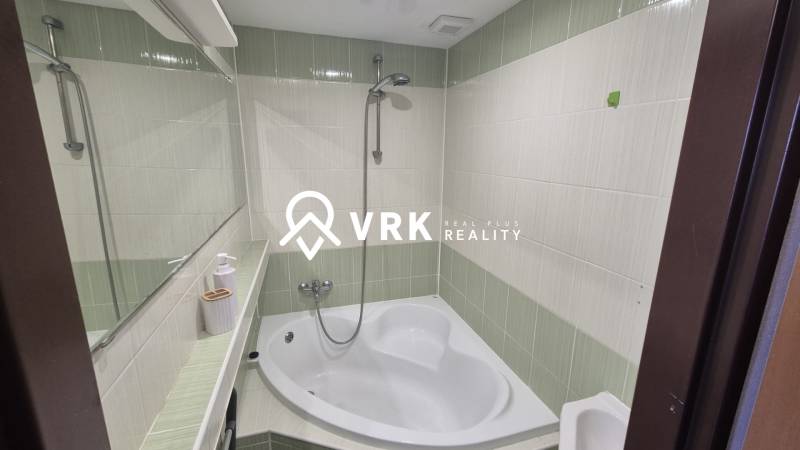 A bathroom with a corner bathtub and green and white tiles in a 2-room apartment.