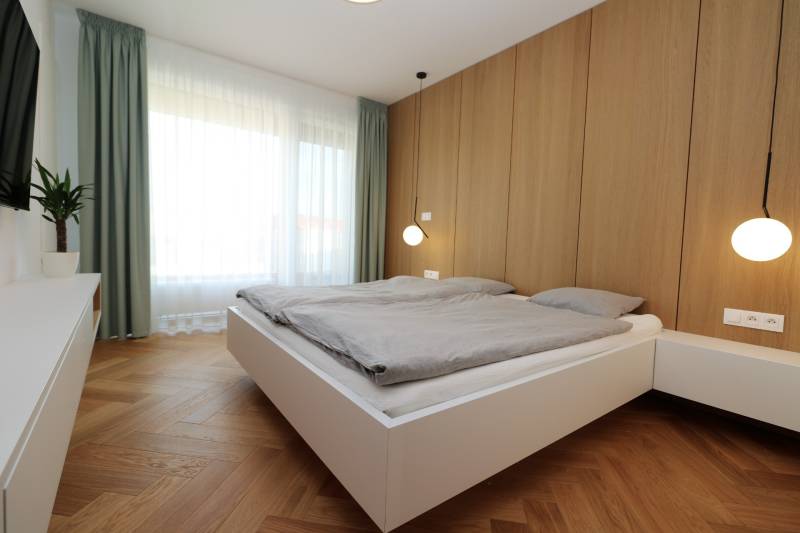 A bedroom in a 4-room apartment with wood-patterned flooring, white furniture, and a large window.