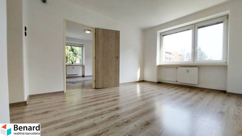 Two-room apartment with wood-patterned flooring, large window, and white walls.