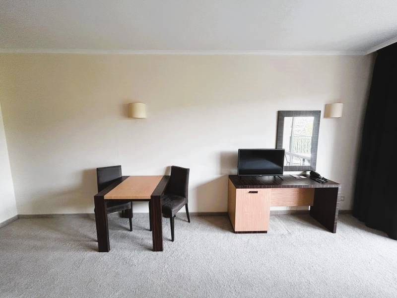 A room with a carpet contains a table, chairs, and a desk with a monitor.