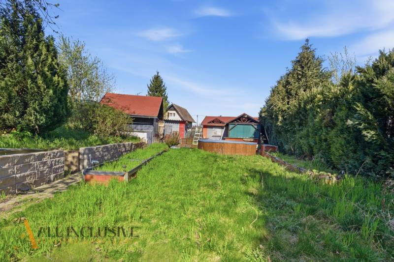 Gardens in Kráľová nad Váhom with a cottage, dense vegetation, and a lawn under the blue sky.