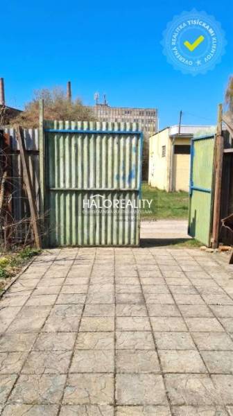 A yard by the cottage in Poltár with a gate and concrete paving under a blue sky.