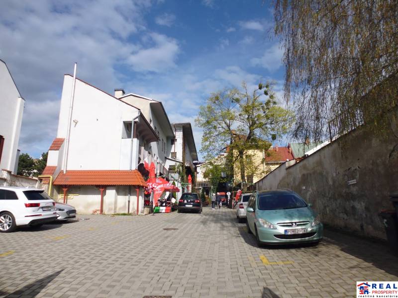 In Prešov on Hlavná Street, there are parked cars and pedestrians on the paved area.