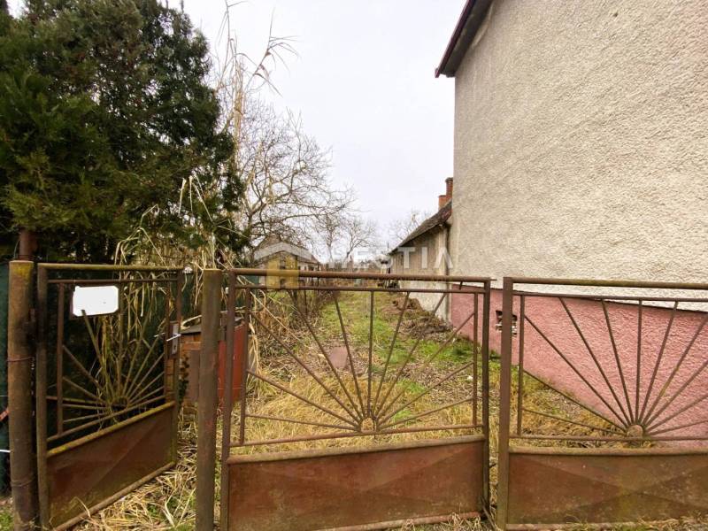 An old fence blocks the entrance to an overgrown plot. A family house with a white facade on the side.