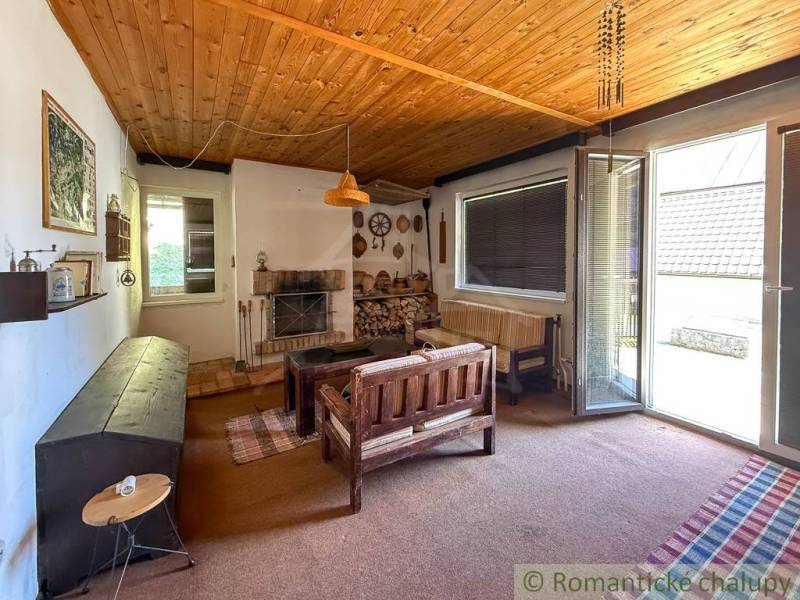 Interior of a cabin with a wooden ceiling, a sofa set, and a fireplace, with a view of the garden.
