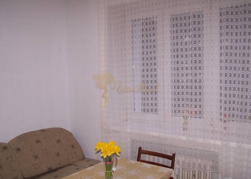 A room with a sofa, table, chair, and curtain, on the table a vase with flowers.