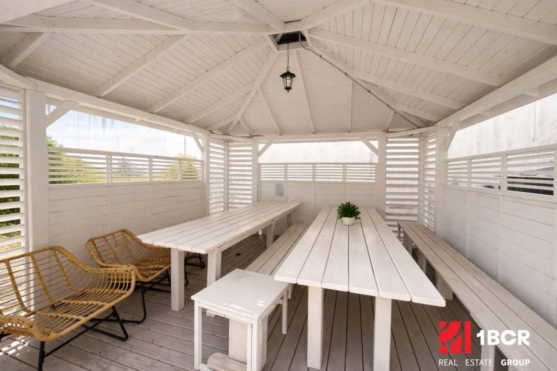 White gazebo with wooden furniture and rattan chairs in the garden.