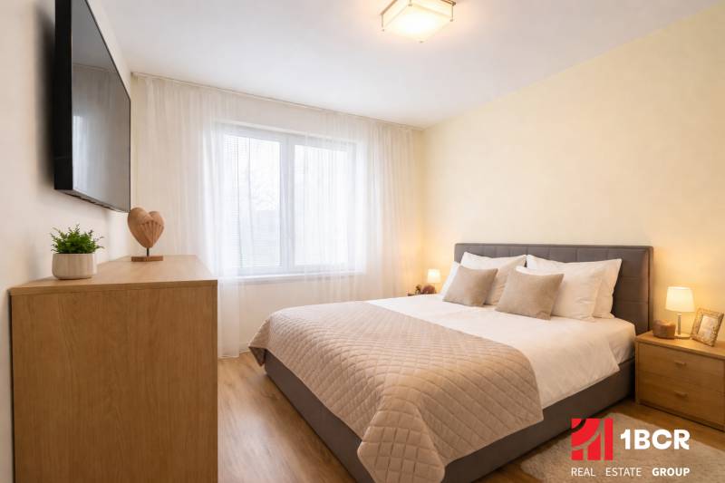A bedroom in a 3-room apartment with a gray bed, a floor with a wood decor, and a TV on the wall.