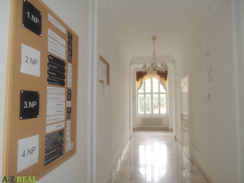 A hallway in offices with a decorative chandelier, white walls, and a board with company names.