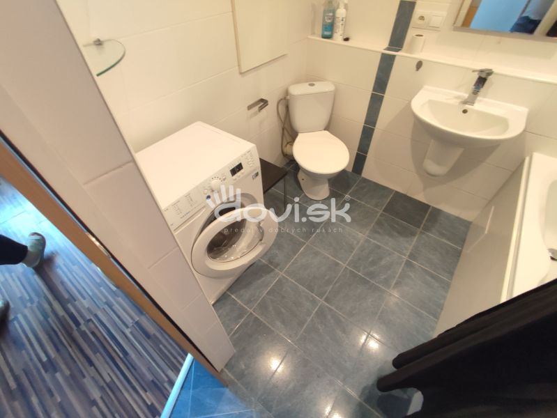 A bathroom with a washing machine, toilet, and sink in a studio apartment with blue tiles.