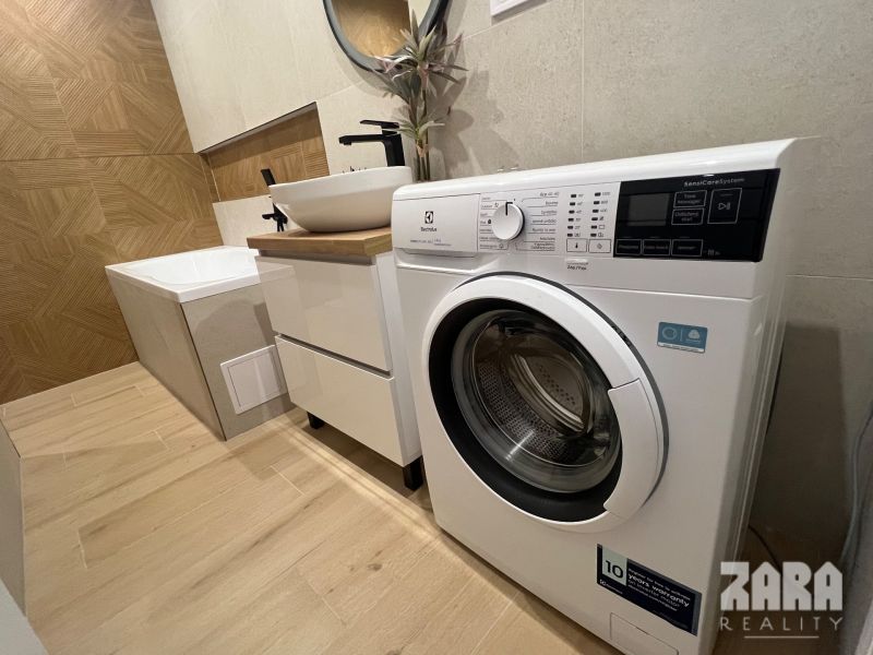 Bathroom in a 2-room apartment with a washing machine, a sink on a cabinet, and a bathtub on a floor with a wooden decor.